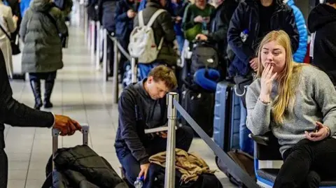 Travelers queue with their luggage at Schiphol Airport. One woman is sitting with her phone in her hand, while a man behind her is kneeling next to his bag while reading a book.