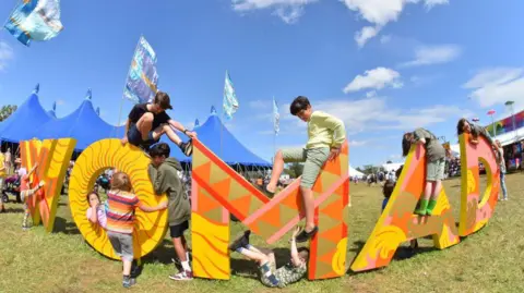 WOMAD sign in orange, yellow, brown and pink. Children are climbing on the letters and there is a blue big top tent in the background.