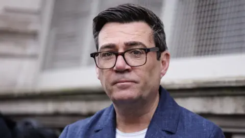 EPA Andy Burnham pictured outside a grey stone building. He wears a royal blue blazer, white T-shirt and black rimmed glasses 