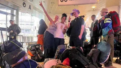 Laurence wears a black T-shirt, black shorts and a purple hat. He stands next to his friends Meg (left) and Kitty (middle) in the coach station surrounded by bags. They smile at the camera as Meg and Kitty do peace signs
