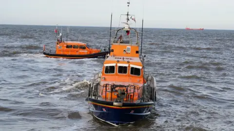 The Trent-class lifeboat Samarbeta at sea off Great Yarmouth. There is another smaller lifeboat behind it. In the distance, on the horizon, there is a cargo ship.