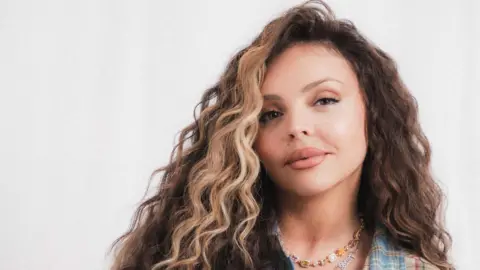 Prime Video Jesy Nelson is looking at the camera. She has brunette curly hair with a blonde streak. She is wearing a necklace that says her daughters names Story and Ocean. 