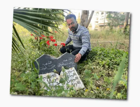 Mohammed wears a striped grey top with a collar, and kneels down next to a black and white grave inscribed with Arabic. There are red flowers planted next to it.