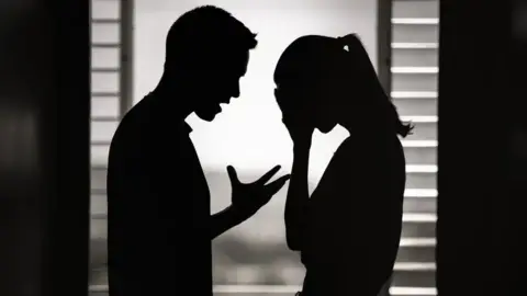 Getty Images File picture of the silhouettes of a man and a woman apparently arguing - the man has the fingers of his right hand stretched out and he is looking at the woman standing close to him, who has her head in her hands. There are closed blinds behind them.