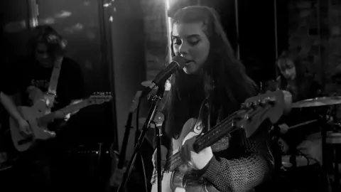 Permanent Daylight A black and white image of Faith Robertson-Charnley playing guitar and singing into a microphone. Behind her Joseph Sullivan plays guitar. 