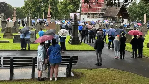 Mourners stand in pairs under umbrellas in a graveyard with their backs to the cameras facing the church in the distance