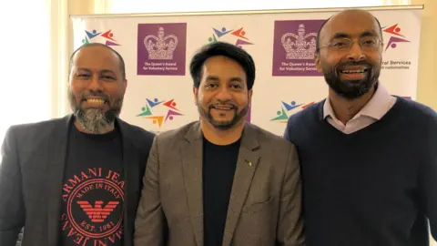 Mohammed Mainul Alam, Boshor Ali and Mojlum Khan stand in front of a branded backdrop which includes the Queen's Award logo