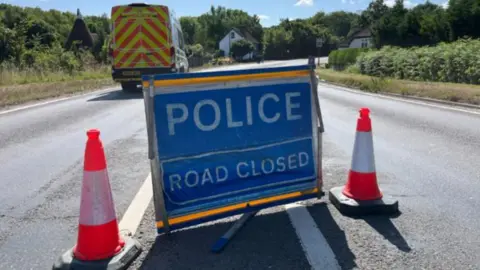 Phil Harrison/BBC Two bollards block off a road, while a blue sign reads 'Police Road Closed' in white letters. A police van is parked in the distance.