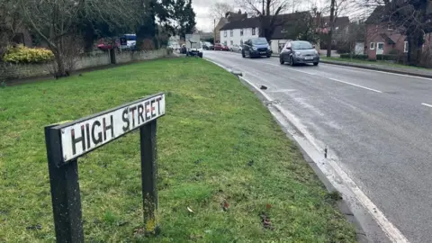 Kate Bradbrook/BBC High Street, Chelveston. A small road lined with trees and several houses. There are cars in the distance and a road sign on the left giving directions for the B645. A High Street sign is to the left by a large area of grass, with a wall in the distance. 