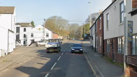 A street with white-fronted houses on both sides and a car and a small truck on the road