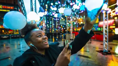 Highcross Shopping Centre A woman holding a hanging light-bulb which is part of an illuminated art installation and taking a picture with her phone.