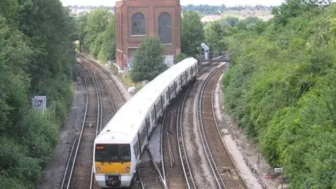 A train is on the track at Dartford Junction, crossing from one side to the other. There is a building where the junction divides into two routes. Trees line the junction on either side.