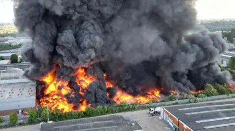 Large area of industrial estate engulfed in fire, with huge clouds of thick black smoke rising in the air. There is a grey warehouse building to the right and further buildings in the foreground with dark grey roofs. 