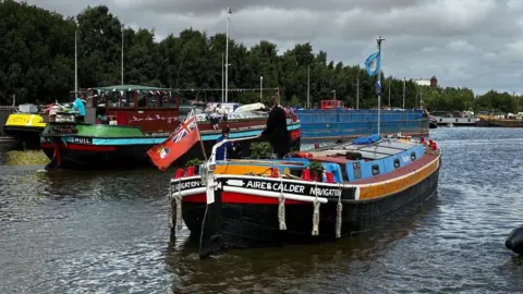A boat with Aire and Calder Navigation on it is in the middle of the waterway. It has two flags on masts. Another barge with Hull written on it is behind it.