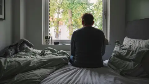 Getty Images A man sits on a bed with green and grey sheets, seen from behind, looking out of a window towards trees outside.