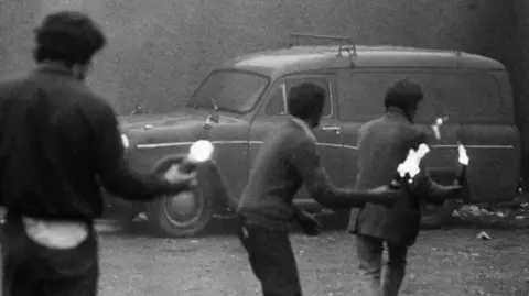 Three young people, throwing petrol bombs towards a car, with their backs facing camera