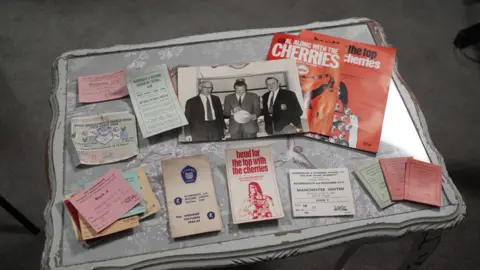 Collection of sports-related memorabilia arranged on a light-colored coffee table. Items include several paper tickets, booklets, leaflets, a black-and-white photograph of three people standing together, and a red magazine with the word “CHERRIES” visible on the cover. The materials are spread across the tabletop in an organised display, with small stacks of tickets and documents positioned around the photograph and magazine. The table sits on a grey carpeted floor, and part of a chair leg is visible at the edge of the image.