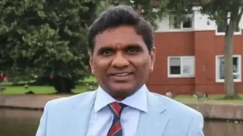 Stoke-on-Trent City Council A man wearing a pale blue suit jacket with a white shirt and a striped tie. He is standing outdoors with trees and a brick building behind him.