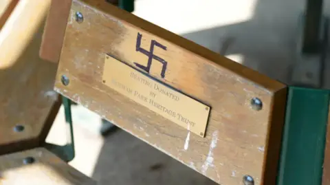 An image of one of the benches in Hotham Park, Bognor Regis, with a swastika drawn onto the wood with what looks like black marker pen, above a plaque which reads "Seating donated by Hotham Park Heritage Trust".