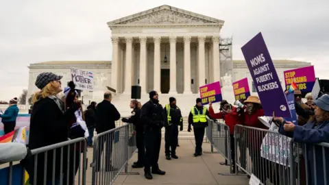 Groups of protestors stand on either side of crowd control barriers outside the Supreme Court building in Washington DC. People hold signs in support of and against gender transition treatment for children. Uniformed police officers stand in an aisle between the two sides