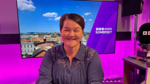 Emma Britton in the BBC Radio Somerset studio. She has short dark hair, is wearing a blue top in a purple radio studio with a screen and a microphone.