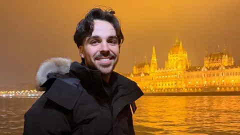 Man with short brown hair and a beard and wearing a black coat is smiling at the camera. Behind him is a river and an ornate building in an orangey/yellow hue.