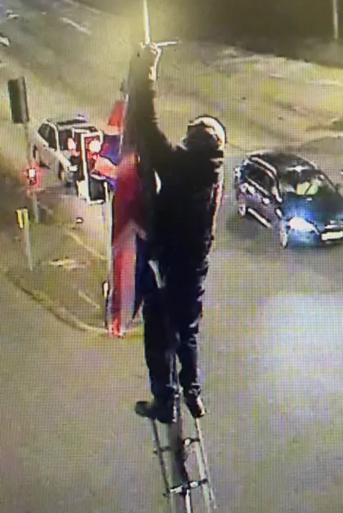 Devon County Council A CCTV image of a man standing on a ladder which is leaning against a lamppost with a Union Flag
