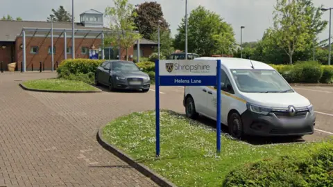 Google Exterior of a brick building and signage on a white background with blue letters saying 'Shropshire County Council' and 'Helena Lane' in white letters with a blue background. A white van is parked next to the sign. A dark coloured car is parked behind the van.