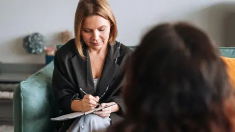 A woman sits in a sofa and makes notes on a clipboard, while a woman (seen from behind) sits opposite her. They are in a room with grey walls and the sofa is green.