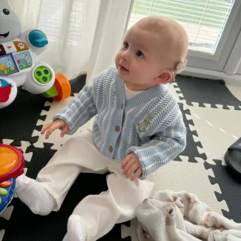 Morgan Deakin A smiling baby wearing a blue cardigan sitting on a black and white play mat