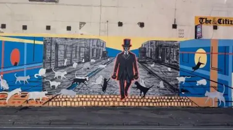 A view of a mural on a building. It shows a man in a black coat and hat surrounded by cats.