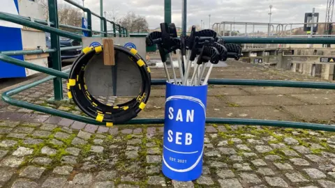 A round blue container - with the label San Seb - full of black-handled litter pickers resting on some cobbles and leaning against a set of green metal railings. To the left are a stack of what appear to be black hoops hung on a wooden peg. Behind them can be seen more railings and the top of a concrete structure forming part of the Tees Barrage