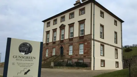 Jennifer Petrie A large cream coloured period house with a floor of sandstone frontage. In the foreground is a sign saying Gunsgreen - House of Secrets