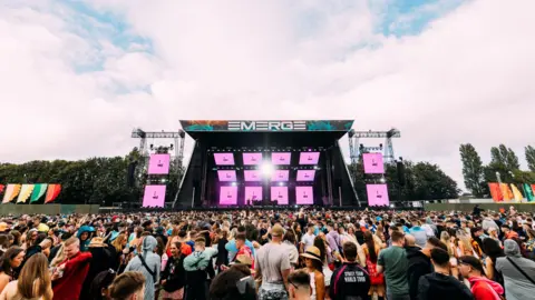 Emerge/The Hype Factory A wide image of a crowd of people attending a large stage show at Belfast Boucher Playing Fields. There are 12 screens arranged on stage, with two stacked vertically on either side of the stage, displaying a pink screen.