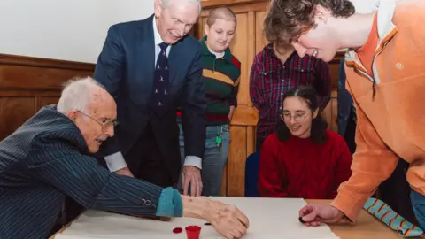 University of Cambridge Bill Steen, far left, with Peter Downes, playing tiddlywinks with current Cambridge University staff and students. They are sat around a wooden table, with a white mat atop, trying to get the circular pieces into a small red cup. 