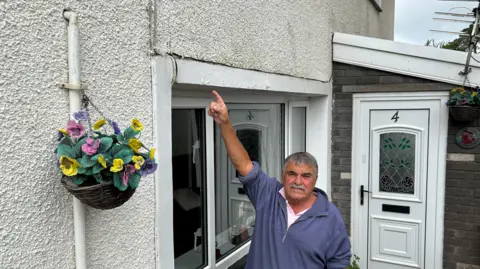 Daryl Wilkins pointing to a crack above his head in the external white render of his home. He is wearing a blue sweatshirt and has grey hair and a grey moustache. He is standing in front of a window, with his front door to the right and a hanging basket to his left.