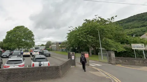 Google A google maps street view image of the school. A pupil is walking through a brick gate on the pavement beside the road into the school There's a car park full of cars and the two storey grey brick school building is in the backgrounds behind some green trees and steep green hill in the distance