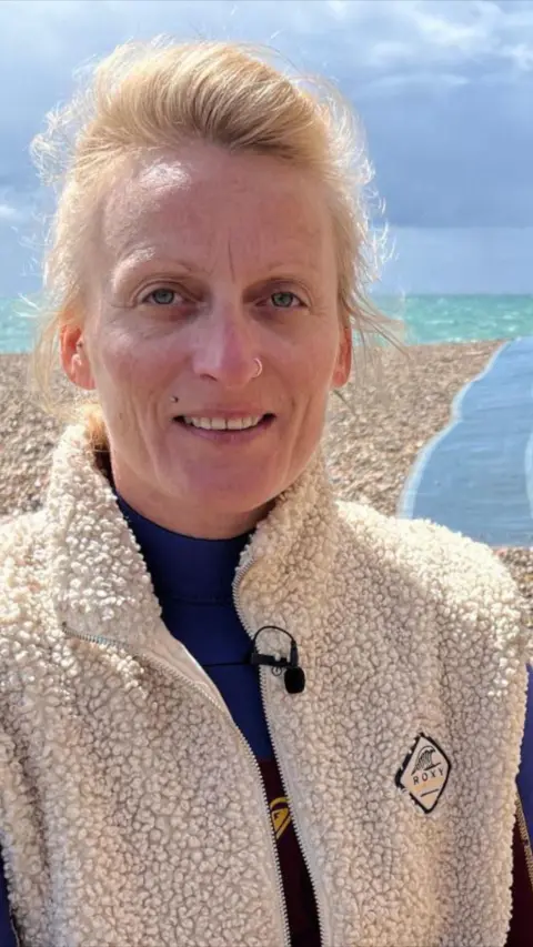 Blonde woman wearing beige fleece standing in front of sea and stoney beach