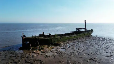 Ministry of Defence A photo of a rusty, old boat which has previously been used for target practice. It is placed on a sandy beach with the sea surrounding it.
