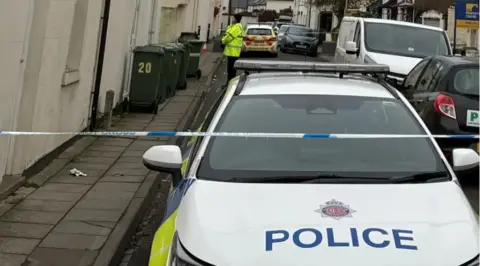 BBC A police car parked close to the camera in the St Pauls area of Cheltenham. In front of it is a line of blue and white police tape stretched across the road. In the background a lone police officer in a high-vis top can be seen, along with some black wheelie bins

