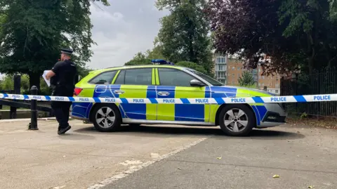 Pete Cooper/BBC A police car is parked across a footpath near a river. A police officer is checking paperwork with his back to the camera. Blue and white police scenes of crime tape has been placed in front of him attached to a metal bollard to the left and iron fencing to the right.
A block of flats is visible in the background on the other side of the river.