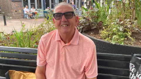LDRS A man with short grey hair, sunglasses and a pink shirt smiles as he sits on a black bench in a town centre. There are several large planters behind him.