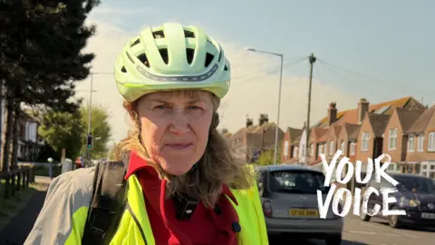 an older woman with long hair, wearing a hi vis jacket and a cycling helmet, looks seriously at the camera as she stands in a street. Cars are seen driving on the road behind her and there are rows of houses on both sides of the road. The words YOUR VOICE are superimposed in white across the right of the picture