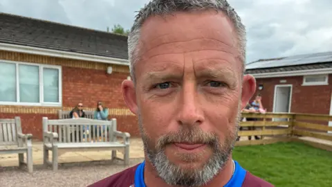 Lewis Moss, with short Grey hair with a black and grey beard. He is in rugby uniform standing in front of several benches