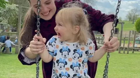 Isabella smiling on a swing with her mother smiling and standing behind her. Isabella has a T-shirt on with Minnie Mouse's face on it.