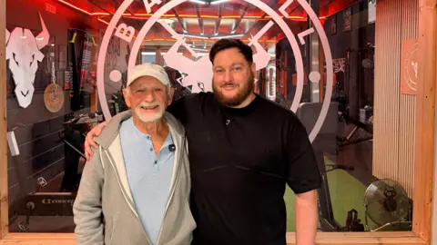 A tall man with dark hair and a beard stands with his arm around an older man with a white beard and moustache, wearing a cap