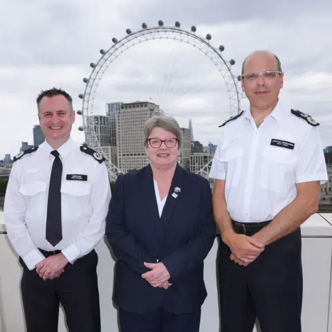 PA Media PC Karen Giles stands alongside Commander Pete Stevens (left) and Assistant Commissioner Matt Twist (right) during a celebration of her 50 years of service at New Scotland Yard in central London. Pc Giles is wearing a dark navy blazer and trousers, with a white blouse underneath and glasses. The two officers beside her are wearing their uniform, which consists of a white shirt with a badge on each shoulder and black trousers. Commander Pete Stevens is also wearing a black tie.