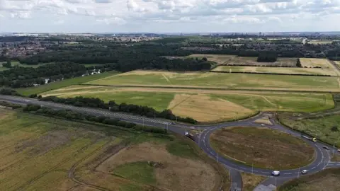Aerial view of the proposed site. There is a large expanse of grassland to the side of a roundabout, with housing in the distance.
