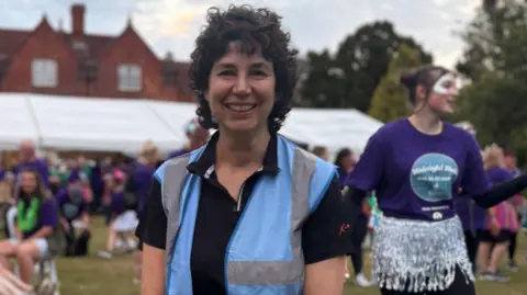 St Luke's Cheshire Hospice A woman with dark curly hair, in a blue hi-viz top over a black polo shirt. standing in front of a marquee, with people behind and to the side of her