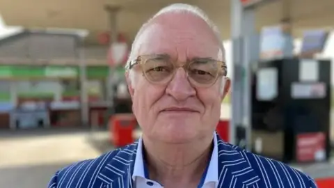 Close up of Ian Mean who is wearing glasses and has short white hair. He is standing in a petrol station wearing a white-striped blue suit.